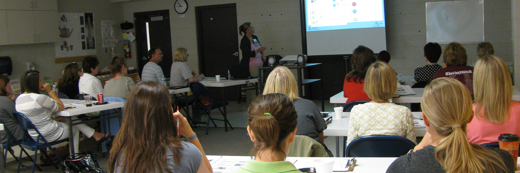 This is an image of an IRCA workshop lecture room. Adult learners at table listening to presenter at front of room pointing to a PowerPoint slide on screen.